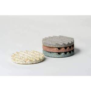 A yellow, grooved, terrazzo concrete coaster is shown beside a stack of three matching ones in grey, red and blue.